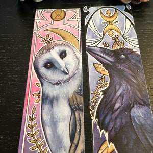 Op de afbeelding: Twee bladwijzers met aquarel illustraties van een uil en een raaf. De uil bladwijzer heeft een roze en paarse achtergrond met gouden accenten. De raaf bladwijzer heeft een blauwe en paarse achtergrond met gouden accenten. Beide bladwijzers bevatten een halve maan en andere geometrische vormen.