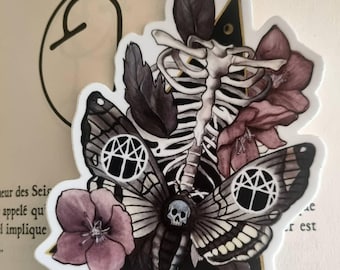 Puede incluir: Una pegatina con una columna vertebral esquelética y una polilla con una calavera, rodeada de flores y hojas de color púrpura oscuro. La polilla tiene intrincados patrones de alas y un diseño geométrico. La pegatina está en una página de libro.