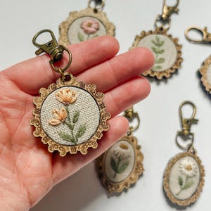 May include: A collection of circular and oval keychains featuring embroidered floral designs on cream-colored fabric. Each piece is encased in an ornate, laser-cut wooden frame with a bronze-toned metal lobster clasp attached to the top.