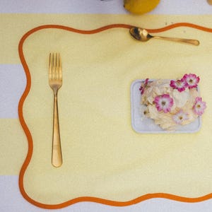 May include: A placemat with a yellow background and an orange border, featuring a gold fork, knife, and spoon. A small plate with a dessert topped with pink flowers is in the center. Lemons are scattered around the placemat.