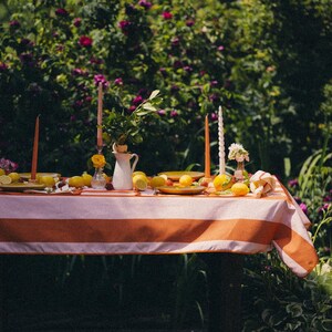 May include: A garden table set with a striped orange and white tablecloth, lemons, candles, a white jug, and small plates. The background shows lush greenery and flowers, creating a summery atmosphere for a gathering.