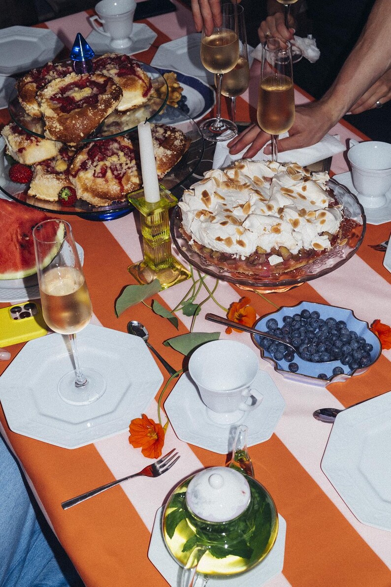May include: A table set for a celebration, featuring a striped orange and white tablecloth. There are pastries, a cake, blueberries, and tea. Champagne flutes and white plates are also present, creating a celebratory atmosphere.