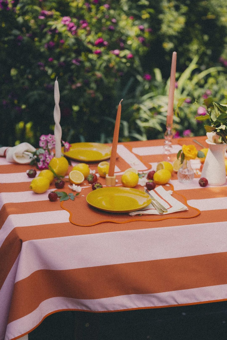 May include: A table set for a meal outdoors, featuring a striped orange and white tablecloth, yellow plates, lemons, strawberries, and candles. The background is a blurred view of flowers. The scene is set for a meal.