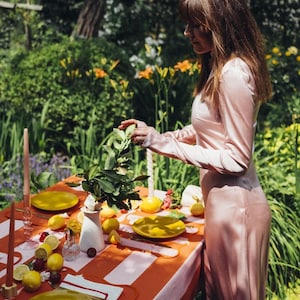 May include: An outdoor table setting with a red and white striped tablecloth. Yellow plates, lemons, and candles are arranged on the table. A person in a pink dress is arranging a floral centerpiece. The scene is set in a garden.