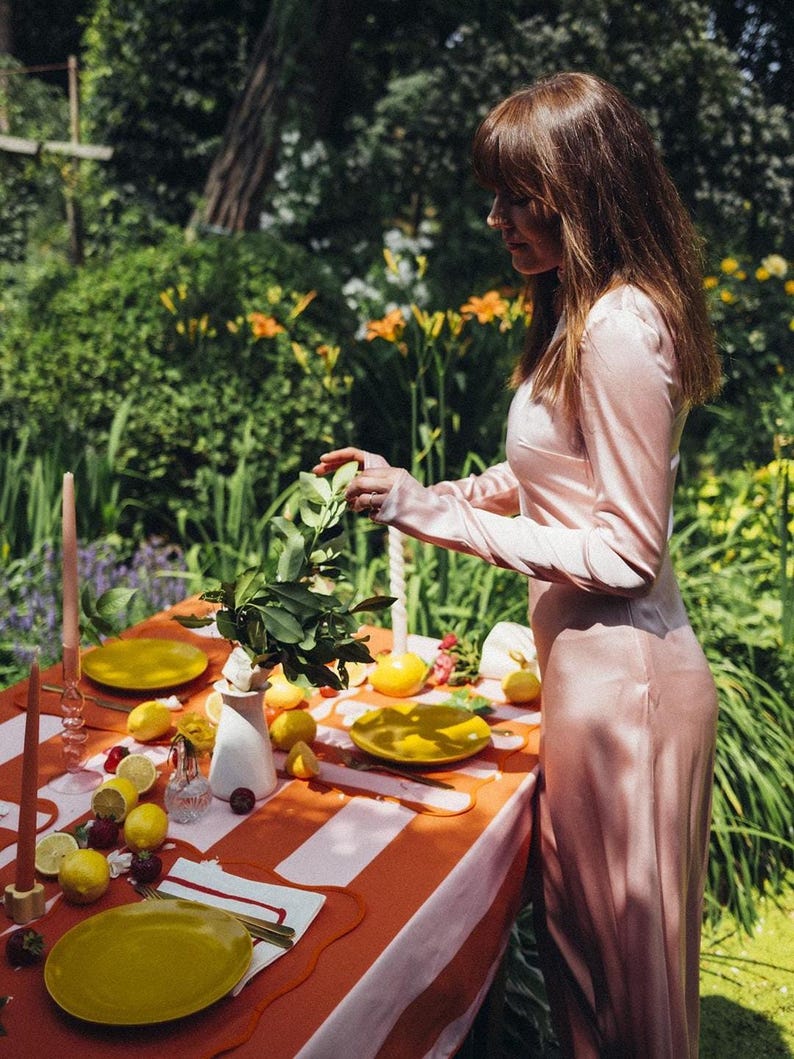 May include: An outdoor table setting with a red and white striped tablecloth. Yellow plates, lemons, and candles are arranged on the table. A person in a pink dress is arranging a floral centerpiece. The scene is set in a garden.