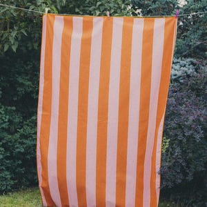 May include: A striped beach towel in shades of orange and pale pink hangs on a washing line. The towel has a vertical stripe pattern and is displayed outside, with green bushes and trees in the background.