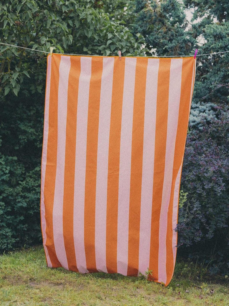 May include: A striped beach towel in shades of orange and pale pink hangs on a washing line. The towel has a vertical stripe pattern and is displayed outside, with green bushes and trees in the background.