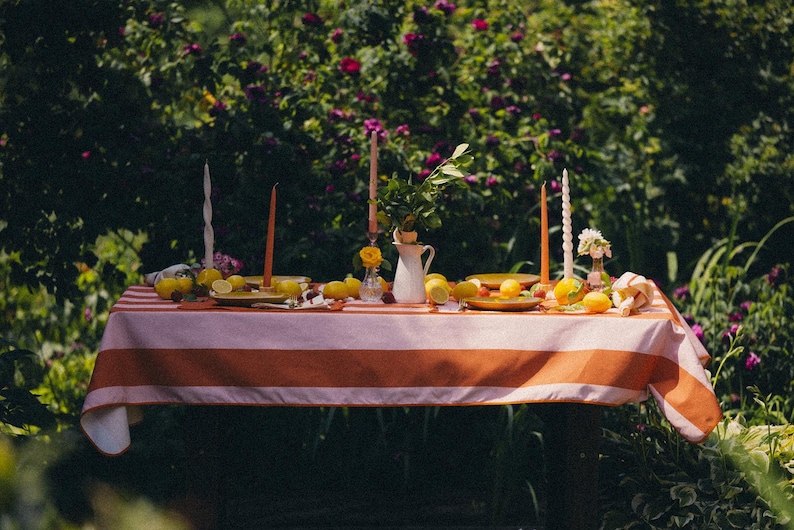 May include: A garden table set with a striped orange and white tablecloth, lemons, candles, a white jug, and small plates. The background shows lush greenery and flowers, creating a summery atmosphere for a gathering.