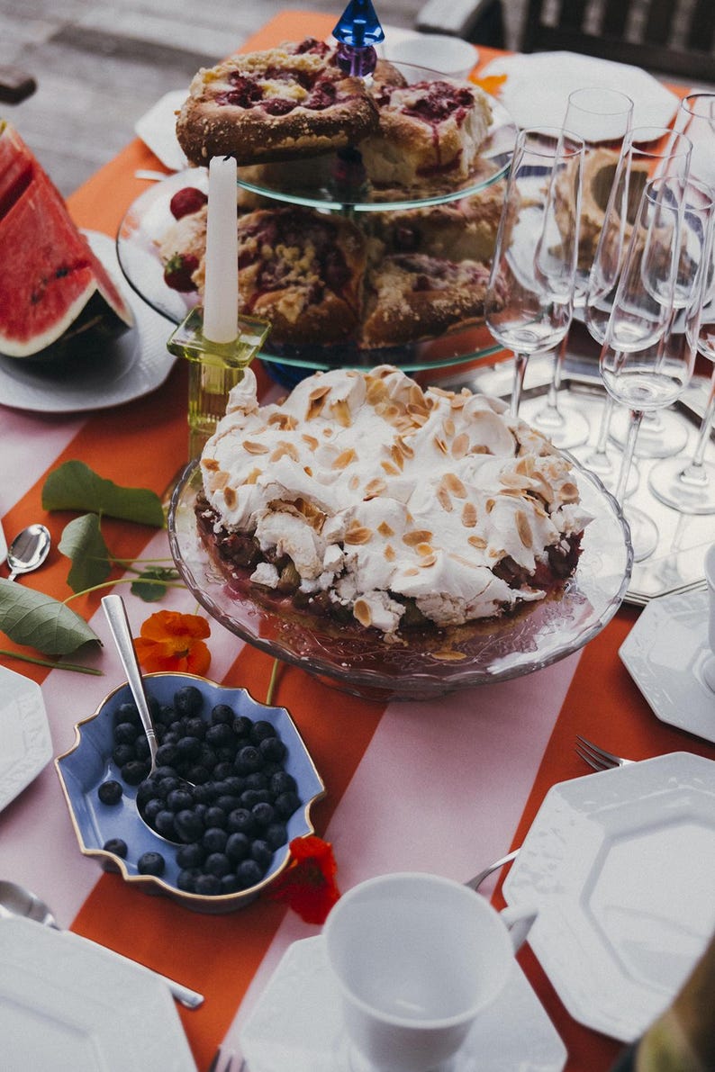 May include: A table laden with treats, including a cake topped with meringue and sliced watermelon. There's a tiered cake stand, blueberries, and champagne glasses. The table is set with a red and white striped tablecloth. The scene suggests a summer gathering.