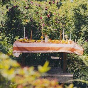 May include: An outdoor table setting with an orange and white striped tablecloth. The table is adorned with lemons, plates, candles, and a small white jug. The background shows abundant greenery and flowering plants.