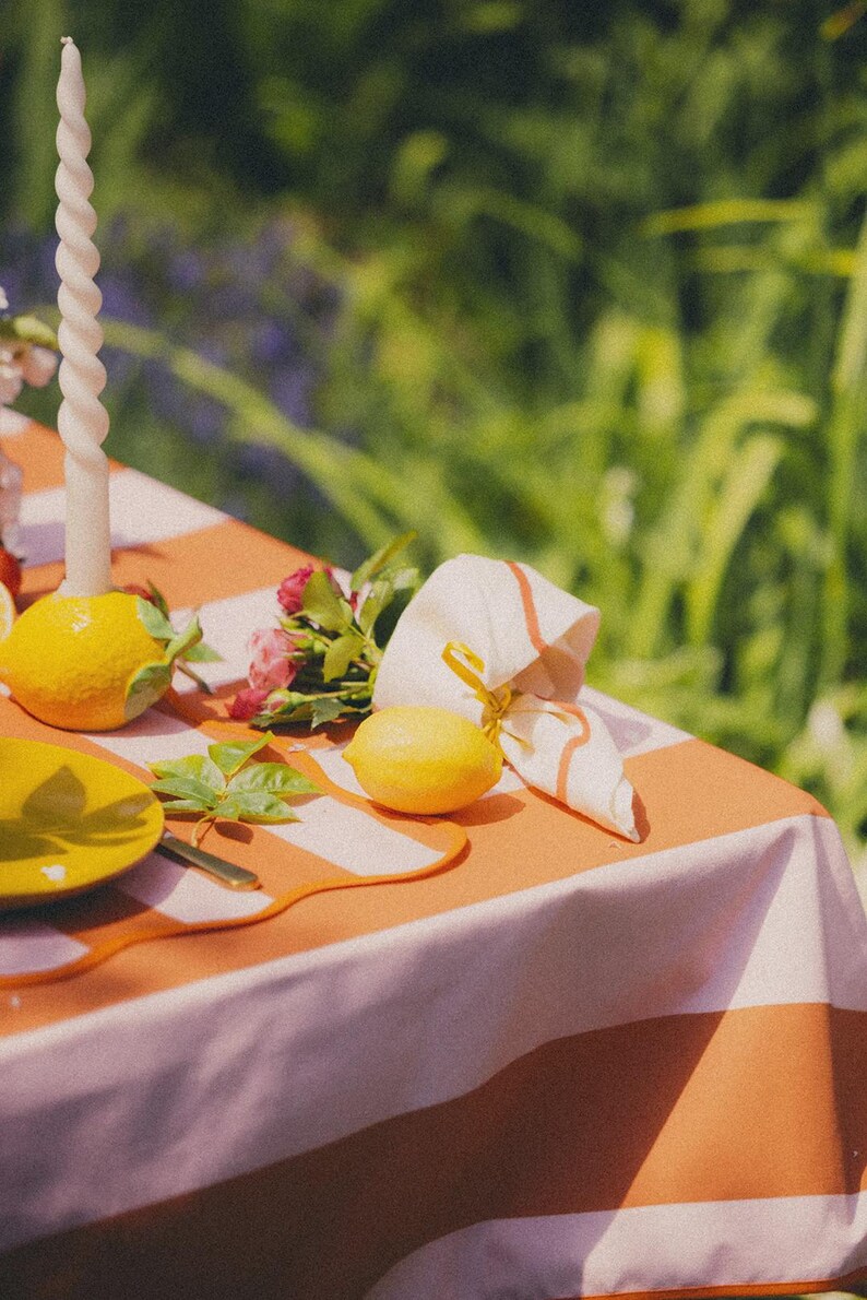 May include: A picnic table set with a white and orange striped tablecloth. The table is decorated with lemons, a yellow plate, a white spiral candle, a napkin, and a small bouquet of pink flowers. The background is blurred green foliage.