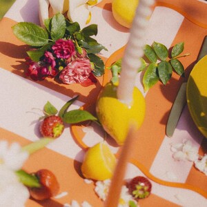 May include: A colourful still life arrangement featuring a striped orange and white surface. The scene includes lemons, strawberries, roses, and a lit candle. The composition evokes a sense of freshness and natural beauty.