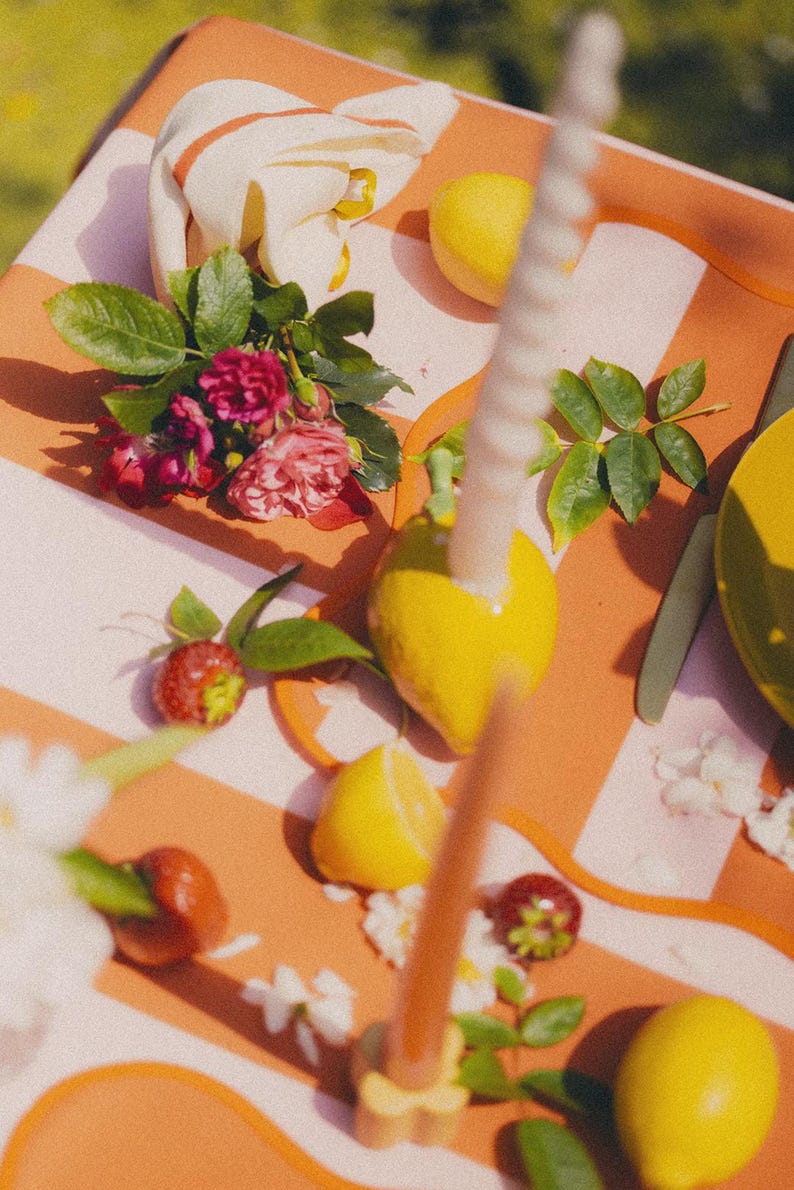 May include: A colourful still life arrangement featuring a striped orange and white surface. The scene includes lemons, strawberries, roses, and a lit candle. The composition evokes a sense of freshness and natural beauty.