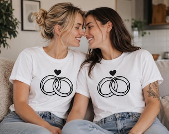 May include: A white short-sleeved t-shirt featuring a black graphic print of two interlocking rings positioned below a small solid black heart. The design is centered on the chest area of the garment, which is made of a plain white fabric.