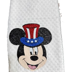 May include: White hand towel with a textured pattern. Features a glittery Mickey Mouse face wearing a red, white, and blue Uncle Sam hat. The towel is a festive and decorative item.
