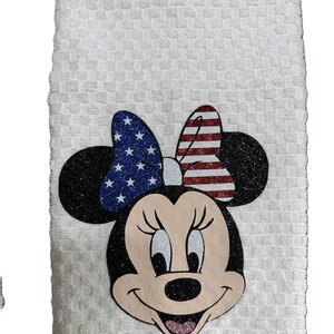 May include: White kitchen towel featuring Minnie Mouse with a patriotic bow. The bow has a blue glitter section with white stars and a red and white striped section. The towel has a textured pattern.