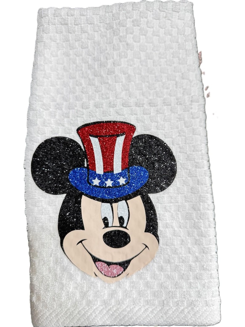 May include: White hand towel with a textured pattern. Features a glittery Mickey Mouse face wearing a red, white, and blue Uncle Sam hat. The towel is a festive and decorative item.