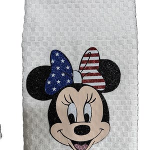 May include: White kitchen towel featuring Minnie Mouse with a patriotic bow. The bow has a blue glitter section with white stars and a red and white striped section. The towel has a textured pattern.