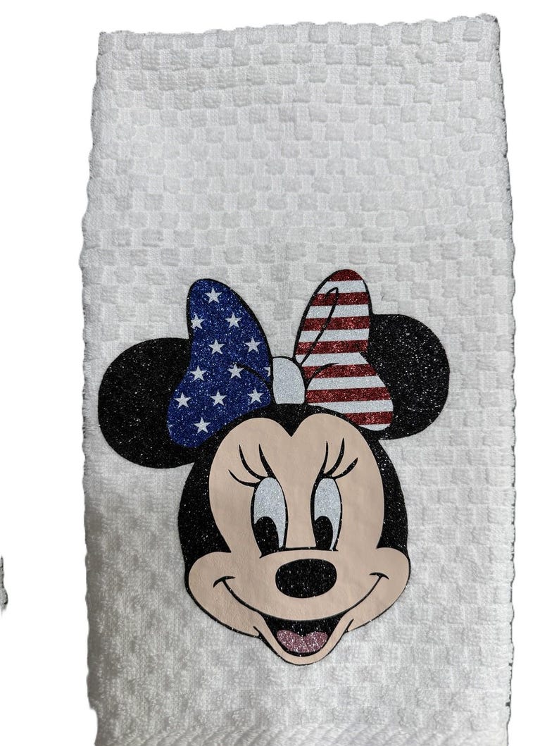 May include: White kitchen towel featuring Minnie Mouse with a patriotic bow. The bow has a blue glitter section with white stars and a red and white striped section. The towel has a textured pattern.