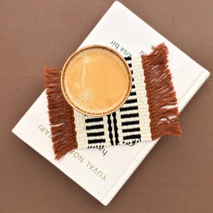May include: A rectangular woven coaster featuring a pattern of cream, black, and rust-coloured threads with fringed edges on two sides. A round ceramic mug filled with a light brown coffee beverage sits centred on top of the woven coaster.