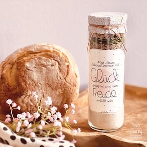 Könnte beinhalten: Ein rustikales Brot liegt neben einem Glas mit Zutaten und einem dekorativen Etikett. Das Glas hat einen Deckel aus gepunktetem Stoff. Das Etikett lautet "Möge dieses Zuhause immer mit Glück gesegnet und von Freude erfüllt sein."