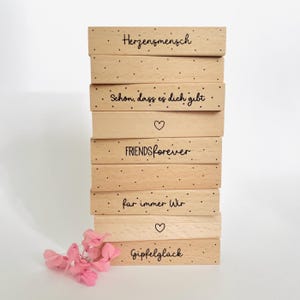 Puede incluir: Una pila de bloques de madera clara con texto negro y pequeños puntos negros. Los bloques tienen frases como "Friends Forever" y "Herzensmensch" impresas. Un pequeño ramo de flores rosas descansa en la base de la pila.
