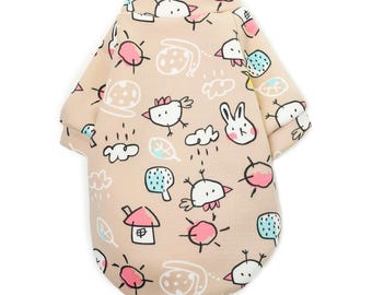 Puede incluir: Un suéter beige para perro con un estampado de dibujos animados de pájaros, conejos, nubes y casas. El suéter tiene cuello redondo y mangas cortas.