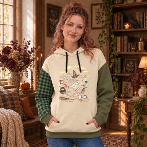 May include: A cream-colored hoodie featuring a front graphic of wizard-themed items including a sorting hat, an owl, a cauldron, and round glasses. The hoodie has a green and black plaid right sleeve, a solid green left sleeve, and black drawstrings.