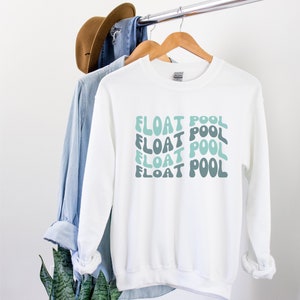 May include: White sweatshirt with the text "FLOAT POOL" repeated four times in a retro, wavy font. The text is in shades of blue and green.