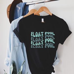 May include: Black t-shirt with the words "FLOAT POOL" in a repeating, wavy design in shades of blue and teal. The shirt is hanging on a wooden hanger with other clothing items in the background.