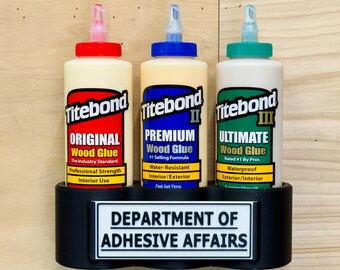 May include: Three bottles of Titebond wood glue in a black holder labeled "DEPARTMENT OF ADHESIVE AFFAIRS." The bottles are labeled "Original," "Premium II," and "Ultimate III." Each bottle has a different colored cap and label.