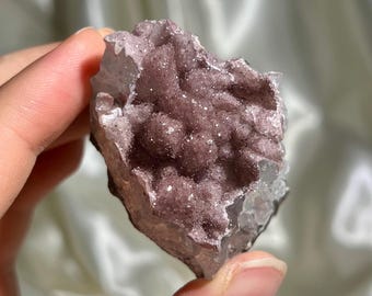 May include: A raw amethyst crystal cluster featuring a deep purple, druzy surface with small, sparkling crystalline formations. The mineral specimen has a jagged, irregular shape with translucent edges and a textured, glittery appearance.