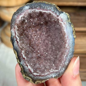 Puede incluir: Una mano sostiene una geoda cortada con un centro de cristal púrpura oscuro. El borde exterior de la geoda es una mezcla de gris, azul y dorado. El interior de cristal brilla con pequeños puntos blancos.