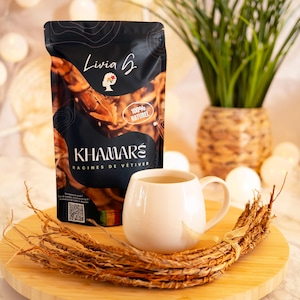 Peut inclure: Une tasse en céramique blanche contenant un liquide brun clair, posée sur une surface en bois avec un paquet de racines brunes séchées. Un sachet noir avec le texte "Livia B. Khamar's Racines de Vetiver 100% Naturel" est visible en arrière-plan.