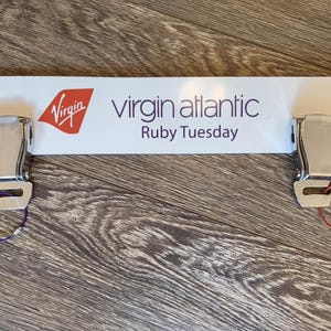 May include: A white sign featuring the Virgin Atlantic logo and the text "Virgin Atlantic Ruby Tuesday." Two silver airplane seat belt buckles are attached, each with a coloured loop: purple and red.