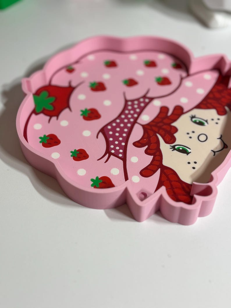 Puede incluir: Una bandeja rosa con forma de fresa con una ilustraci&oacute;n de dibujos animados de una ni&ntilde;a con cabello rojo y ojos verdes. La superficie de la bandeja presenta lunares blancos y fresas rojas.