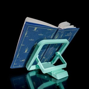 May include: A teal book stand supporting an open book with a blue cover. The book has gold lettering and illustrations of plants. The stand is made of plastic and has an adjustable design.
