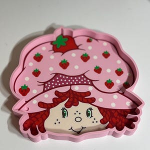 Puede incluir: Una bandeja rosa con la forma de Strawberry Shortcake. La bandeja presenta una cara sonriente, pelo rojo y un sombrero rosa adornado con fresas y lunares blancos. Un art&iacute;culo nost&aacute;lgico.