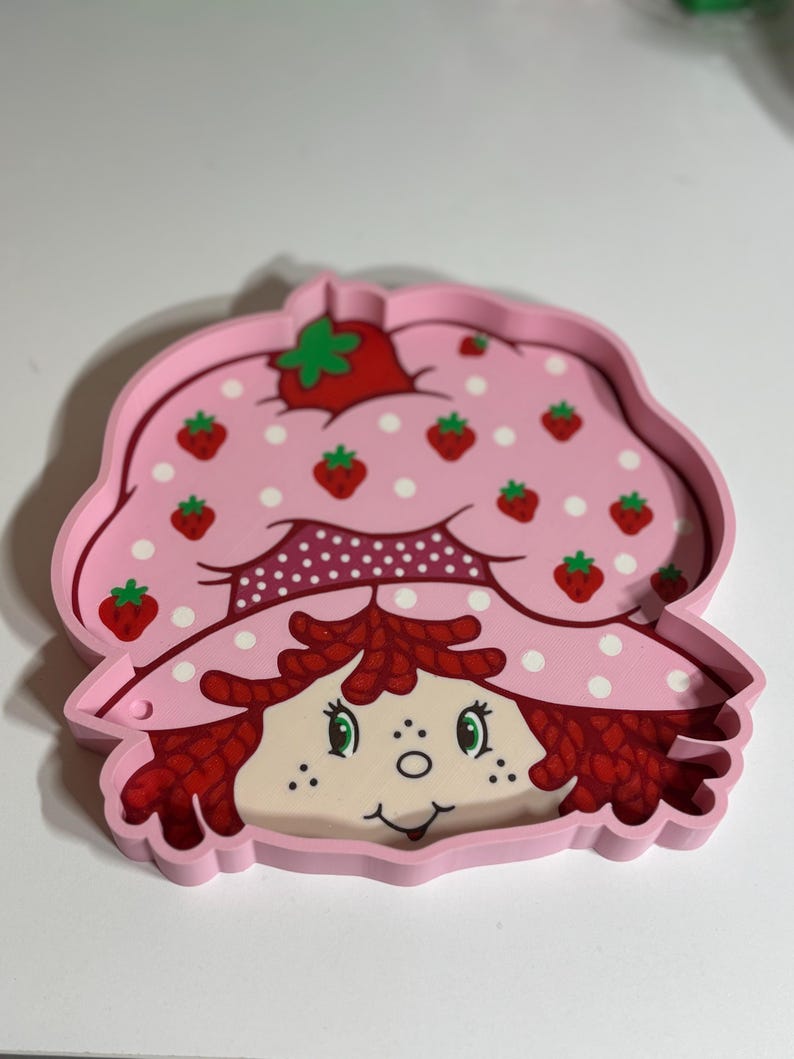Puede incluir: Una bandeja rosa con la forma de Strawberry Shortcake. La bandeja presenta una cara sonriente, pelo rojo y un sombrero rosa adornado con fresas y lunares blancos. Un art&iacute;culo nost&aacute;lgico.