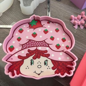 Puede incluir: Una bandeja rosa con tem&aacute;tica de Strawberry Shortcake con una cara sonriente, cabello rojo y un sombrero adornado con fresas. La bandeja est&aacute; decorada con fresas y lunares blancos. Se ven otros moldes y accesorios de artesan&iacute;a.