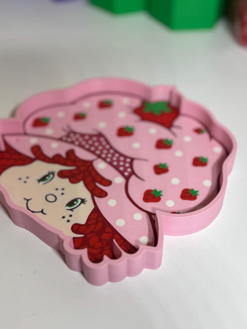 Puede incluir: Una bandeja rosa con forma de fresa con un personaje de dibujos animados. El interior de la bandeja tiene un estampado de lunares rosas y fresas rojas. La bandeja est&aacute; hecha de un material s&oacute;lido y es un objeto decorativo encantador.