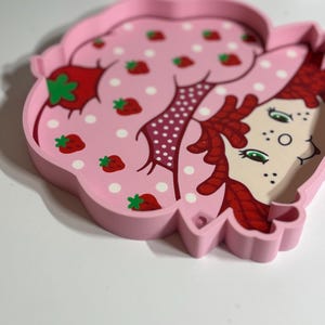Puede incluir: Una bandeja rosa con forma de fresa con una ilustraci&oacute;n de dibujos animados de una ni&ntilde;a con cabello rojo y ojos verdes. La superficie de la bandeja presenta lunares blancos y fresas rojas con hojas verdes.