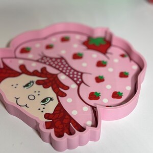 Puede incluir: Una bandeja rosa con forma de fresa con una ilustraci&oacute;n de dibujos animados de un personaje con cabello rojo y un vestido con estampado de fresas. La bandeja tiene un borde elevado y est&aacute; decorada con fresas y lunares blancos.