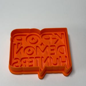 May include: An orange cookie cutter with the text "KPOP DEMON HUNTERS" in a speech bubble design. The letters are in a bold, sans-serif font. The cutter is made of plastic and sits on a white surface.
