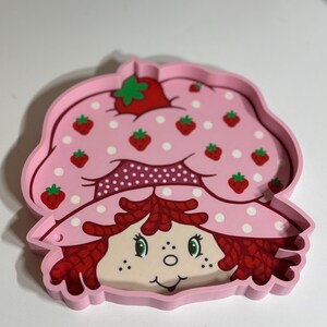 Puede incluir: Una bandeja rosa con la forma de Strawberry Shortcake. La bandeja presenta una cara sonriente, pelo rojo y un sombrero rosa adornado con fresas y lunares blancos. Un art&iacute;culo nost&aacute;lgico para los fans del personaje.