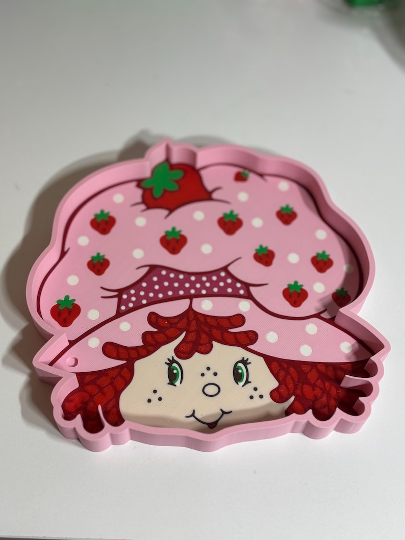 Puede incluir: Una bandeja rosa con la forma de Strawberry Shortcake. La bandeja presenta una cara sonriente, pelo rojo y un sombrero rosa adornado con fresas y lunares blancos. Un art&iacute;culo nost&aacute;lgico para los fans del personaje.