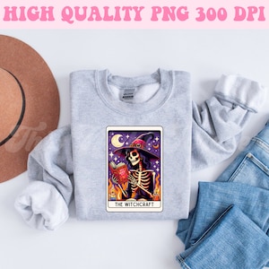 K&ouml;nnte beinhalten: Ein graues Sweatshirt mit einem Tarotkarten-Design, das ein Skelett zeigt, das einen Hexenhut tr&auml;gt und ein Buch h&auml;lt. Die Karte lautet "The Witchcraft".