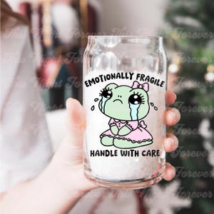 May include: A clear glass can with a green frog wearing a pink dress and a bow. The frog has large eyes and is crying. The text on the can reads "Emotionally Fragile Handle With Care".