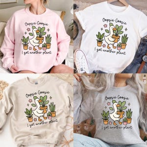 以下が含まれることがあります： 「Oopsie Goosie」と「I got another plant」の文字が入った4種類のトップス。ピンクのスウェットシャツ、白のTシャツ、ベージュのスウェットシャツ、グレーのスウェットシャツがあります。各トップスには、鉢植えの植物を持っている漫画のアヒルが描かれています。