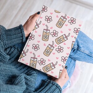 May include: A notebook with a pink cover featuring a repeating pattern of smiling iced coffee drinks, pink flowers, and hearts. The notebook is held open, revealing the patterned design. The overall theme is a cute, whimsical design.